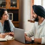 Indian male in turban and wristwatch typing on netbook while interacting with happy female coworker with notebook and pen sitting at table in office and looking at each other